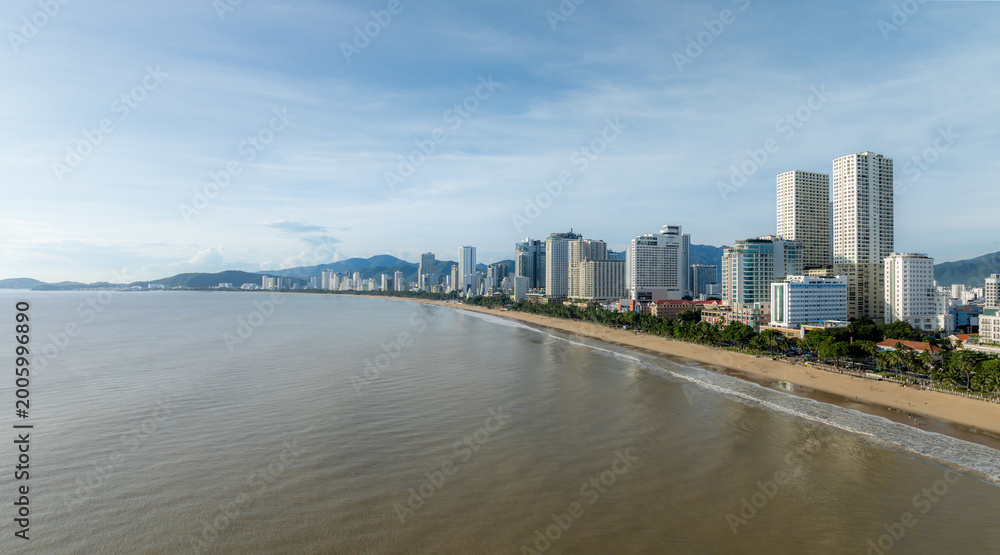 Fototapeta premium A panoramic view of the Nha Trang beach in Vietnam, with its sandy shore, palm trees, and modern high-rise buildings lining the coast. The sea is calm under a partly cloudy sky.