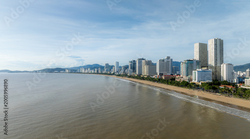 A panoramic view of the Nha Trang beach in Vietnam, with its sandy shore, palm trees, and modern high-rise buildings lining the coast. The sea is calm under a partly cloudy sky.