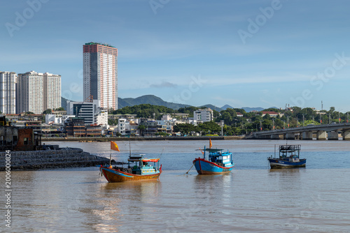 Three fishing boats float on the Cai River, Nha Trang,  Vietnam, with the city skyline and mountains in the background.