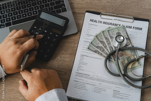 A healthcare professional uses a calculator to carefully process health insurance claim reimbursement costs, with a stethoscope and US dollar bills placed on a medical claim form clipboard.