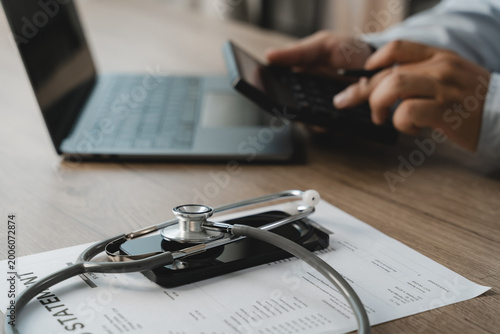 A doctor carefully uses a calculator and laptop to review a medical billing statement for insurance cost analysis, with a stethoscope and smartphone resting on the document on a wooden office desk.