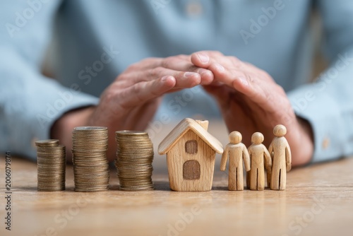Hands shield wooden family, house, and stacks of coins