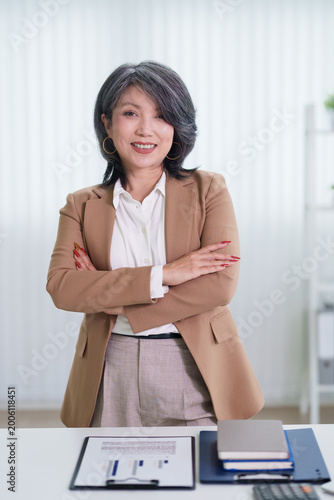 confident mature Asian businesswoman executive smiling arms crossed standing office desk professional corporate workplace bright modern business leader success