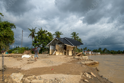 A man walks toward a severely damaged house on riverbank after flash flood in West Sumatra, Indonesia, highlighting erosion, destruction and impact of extreme weather on rural community