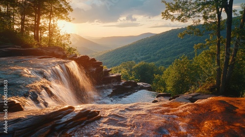 Waterfall edge overlooks a vast valley filled with lush green mountains during a warm and golden sunset hour.