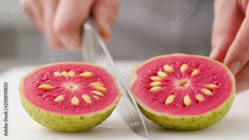 Close-up of a hand cutting a fresh guava in half on a white cutting board.