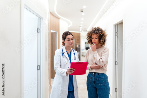 Fotografie Young female doctor showing medical results on a pink clipboard to a pensive patient in a clinic hallway