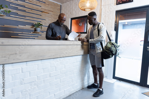 Mature African American men at gym desk reviewing forms, one in gear signing, carrying green duffel