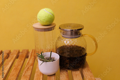 A teapot rests on a wooden table beside a glass jar and a small plant with a tennis ball