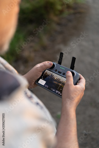 Close-up over-the-shoulder shot of a drone pilot's hands gripping the controller. The bright screen displays live aerial footage, with thumbnails and flight data visible, while thumbs hover over joyst