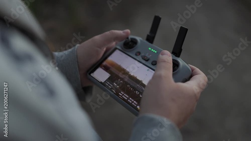 Close-up over-the-shoulder shot of a drone pilot's hands gripping the controller. The bright screen displays live aerial footage, with thumbnails and flight data visible, while thumbs hover over joyst