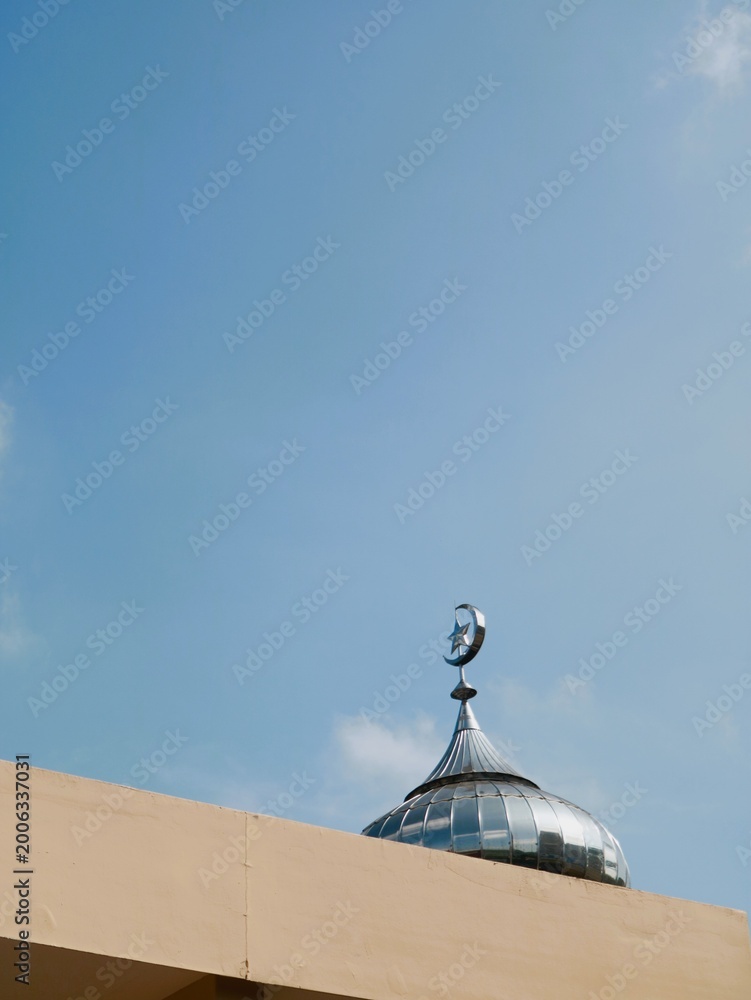 Naklejka premium A silver dome with a crescent moon finial atop a building against a clear blue sky, symbolizing Islamic architecture and faith.