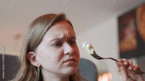 Caucasian woman grimacing at forkful salad, closeup reaction in intimate bistro setting, inspecting greens and dressing with visible freckles and concern, reluctant taste test under soft ambient light