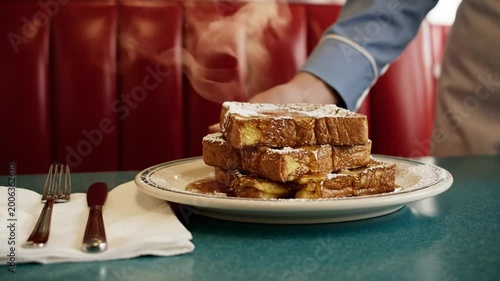 Hot French toast stack with syrup being served in a diner by a waiter in front of red booth seating