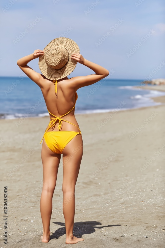 Fototapeta premium Woman adjusting straw hat while standing on wide sandy beach