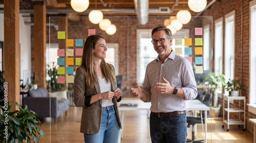 caucasian colleagues brainstorming behind glass board in modern office. smiling man and woman discussing creative project with sticky notes. teamwork, startup concept. banner, website header.