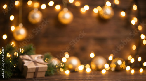 A festive Christmas scene with glowing golden lights and ornaments adorning a rustic wooden background, featuring a gift-wrapped present and evergreen branches.