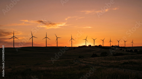 Wide panorama of a wind farm silhouette at sunset, row of wind turbines on rolling hills against a vibrant orange sky.