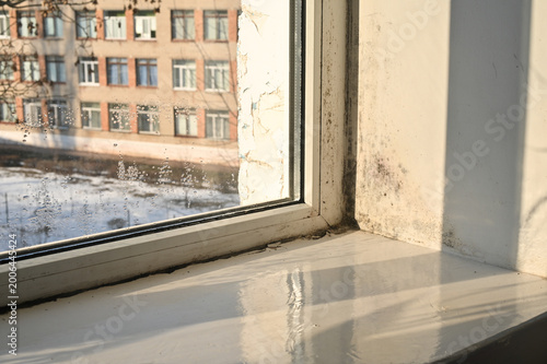 Black mold growing on window slopes and frame due to condensation and high humidity. Mold damaged white wall surface near window. Problem with dampness and mold inside house room.
