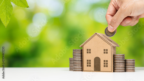 Hand placing coin into house model with stacked coins, home investment concept. Home savings and property investment with coin deposit