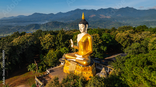 A majestic golden Buddha statue surrounded by lush green trees, set against a stunning backdrop of rolling mountains and a clear blue sky. This serene landscape captures the harmony of spirituality