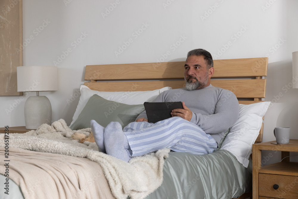 Fototapeta premium Mature man sitting on bed wearing pajama pants, holding tablet and looking right