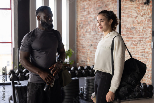 Diverse gym partners in sportswear talking, holding water bottle, duffel bag at gym with dumbbells