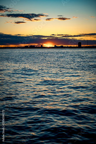 Golden sun setting over ocean with urban skyline silhouette. Atmospheric sunset view with the sun low on the horizon over water and city buildings in distance.