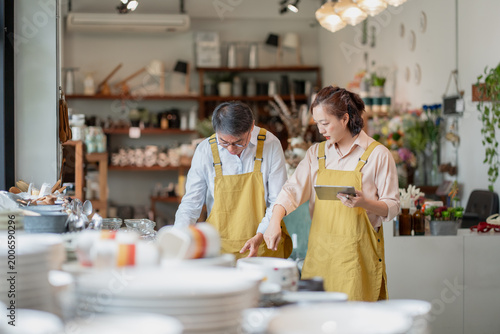 Professional Asian man and woman in aprons working together while checking inventory on a digital tablet in a bright retail store, showcasing teamwork and small business success.
