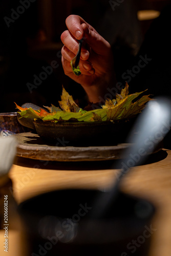 Canvastavla Gros plan sur une main utilisant une pince de précision pour composer un plat gastronomique sur un lit de feuilles d'automne, dans une ambiance tamisée et minimaliste au style raffiné