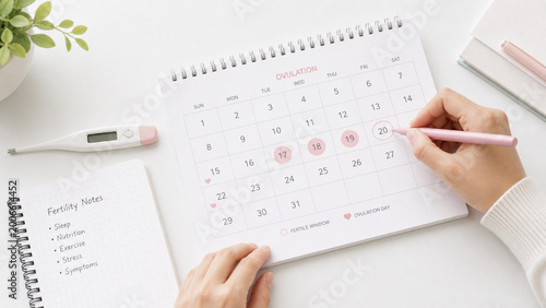 Close-up of hands marking ovulation dates on a calendar with a pen, alongside a thermometer and fertility notes on a clean desk. Concept of fertility tracking, reproductive health awareness