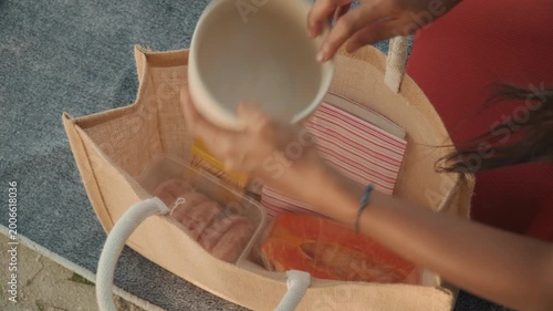 Straw tote packed with picnic essentials, woman reaches into bag to retrieve sealed container and snacks, closeup of hands organizing compartments, red dress sleeve visible, casual beach preparation,