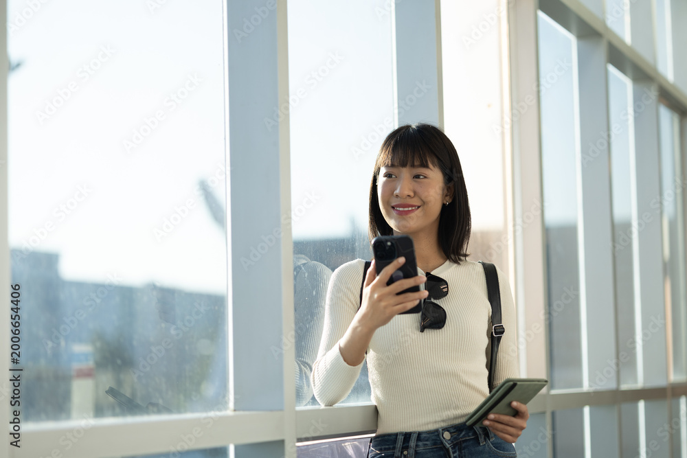 Fototapeta premium Smiling young Asian traveler holding smartphone and passport at airport terminal 