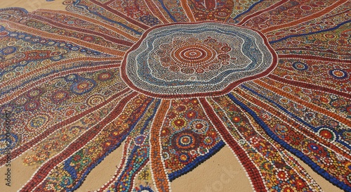 Colorful mosaic artwork on a sandy surface, featuring intricate patterns and radiating designs.  Geometric shapes create a dynamic visual