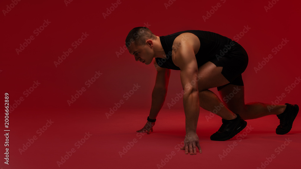 Fototapeta premium Athletic man crouched in explosive sprint starting position against red studio background
