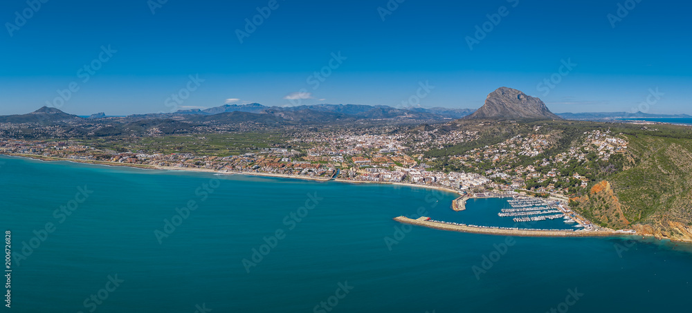 Fototapeta premium Panoramic aerial view of Javea, Costa Blanca, Spain, showing turquoise Mediterranean sea, marina with boats, white town buildings, and iconic Montgo mountain backdrop