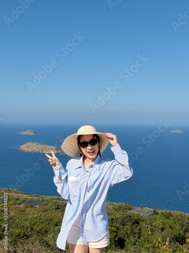Fotografia Young Asian woman stands on hilltop near ocean, wearing a sun hat and smiling wi