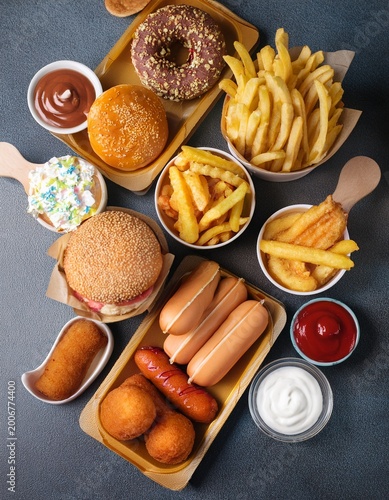 Ultra processed foods. Top view of many glazed donuts, icecreams, fried potatoes, hot dogs, sauces on gray table