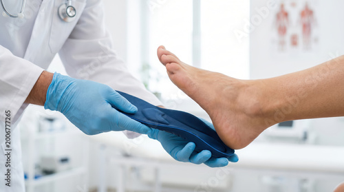 A doctor is fitting a custom foot insole on a patient's foot. The setting is a clinic where medical evaluations take place. The doctor wears gloves and focuses on the patient's foot