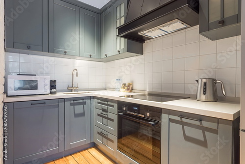 A kitchen features gray cabinets, a stainless steel stove, and a sink. There is a microwave and kettle on the counter. The floor is made of wood. Light comes through a window