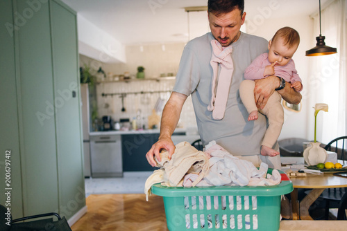 Quadro su tela Busy dad talking on the phone carrying laundry basket and baby