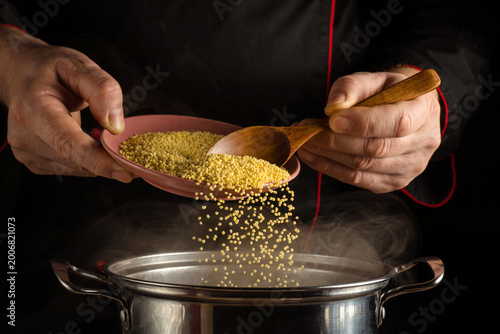Hands are seen pouring millet from a plate into a pot. Steam rises from the pot as food is being prepared. The scene is focused on the activity of cooking