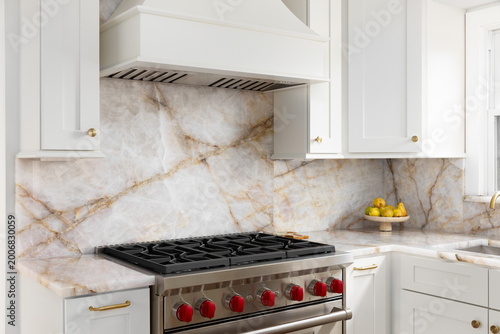 A white kitchen range hood detail with a marble countertop and backsplash, stainless steel oven, and gold accents.
