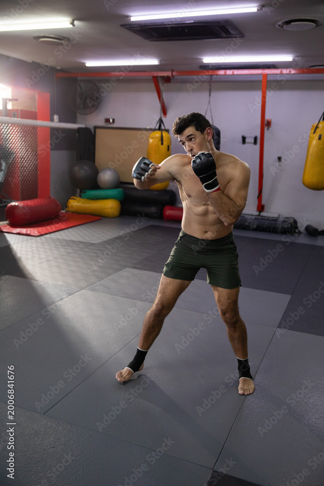 Fototapeta premium Practicing shirtless fighter wearing green shorts, wraps striking in MMA gym with cage and bags