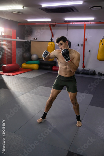 Practicing shirtless fighter wearing green shorts, wraps striking in MMA gym with cage and bags