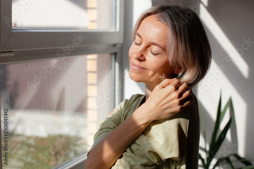 Fotografía Attractive Woman hugs herself, woman standing near window with close eyes at hom