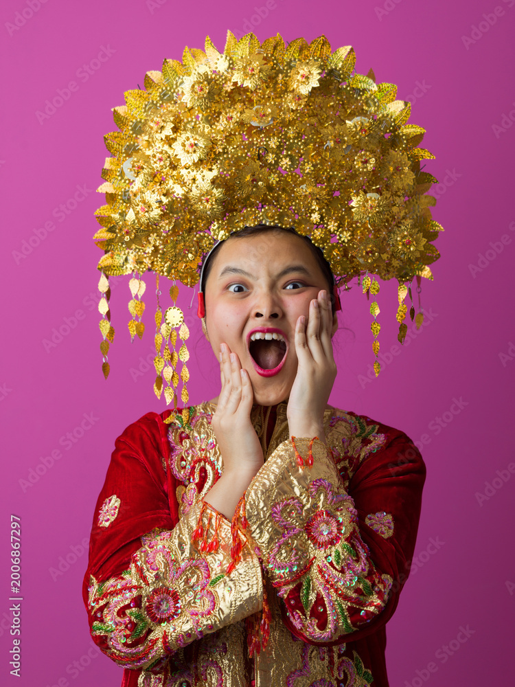 Naklejka premium Surprised Young Indonesian Asian Girl Wearing Ornate Gold Headdress And Embroidered Traditional Costume On Pink