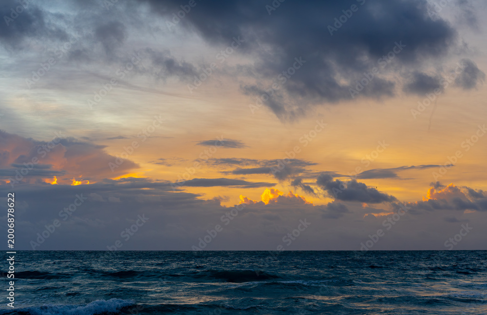 Fototapeta premium Sunset over the ocean with dramatic clouds in Miami. Dark sea waves under a colorful sky with orange light breaking through gray clouds at dusk in Miami, Florida.