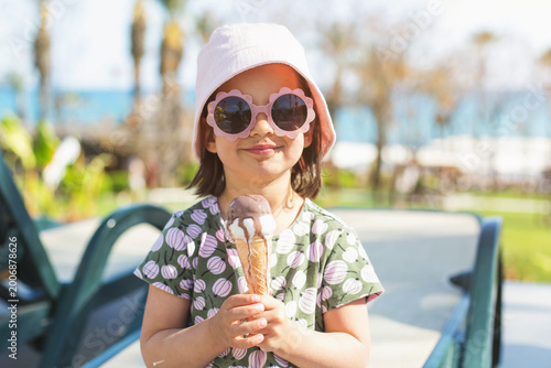 Bild auf Leinwand Happy little girl holding ice cream in sun hat by seaside resort, child in flowe