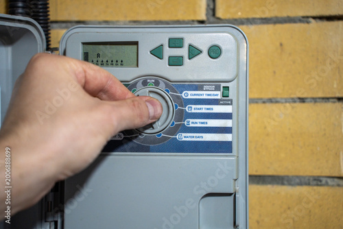Fotografía Hand turning selector dial on irrigation controller panel, adjusting automatic watering system settings on a brick wall